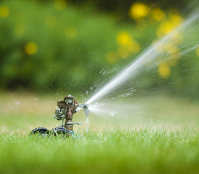 A metal lawn sprinkler spraying a stream of water over green grass with a blurred garden background.