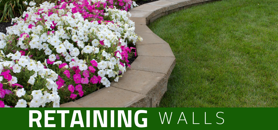A low retaining wall made of concrete blocks curving along a garden bed filled with white and pink flowers next to a lawn.