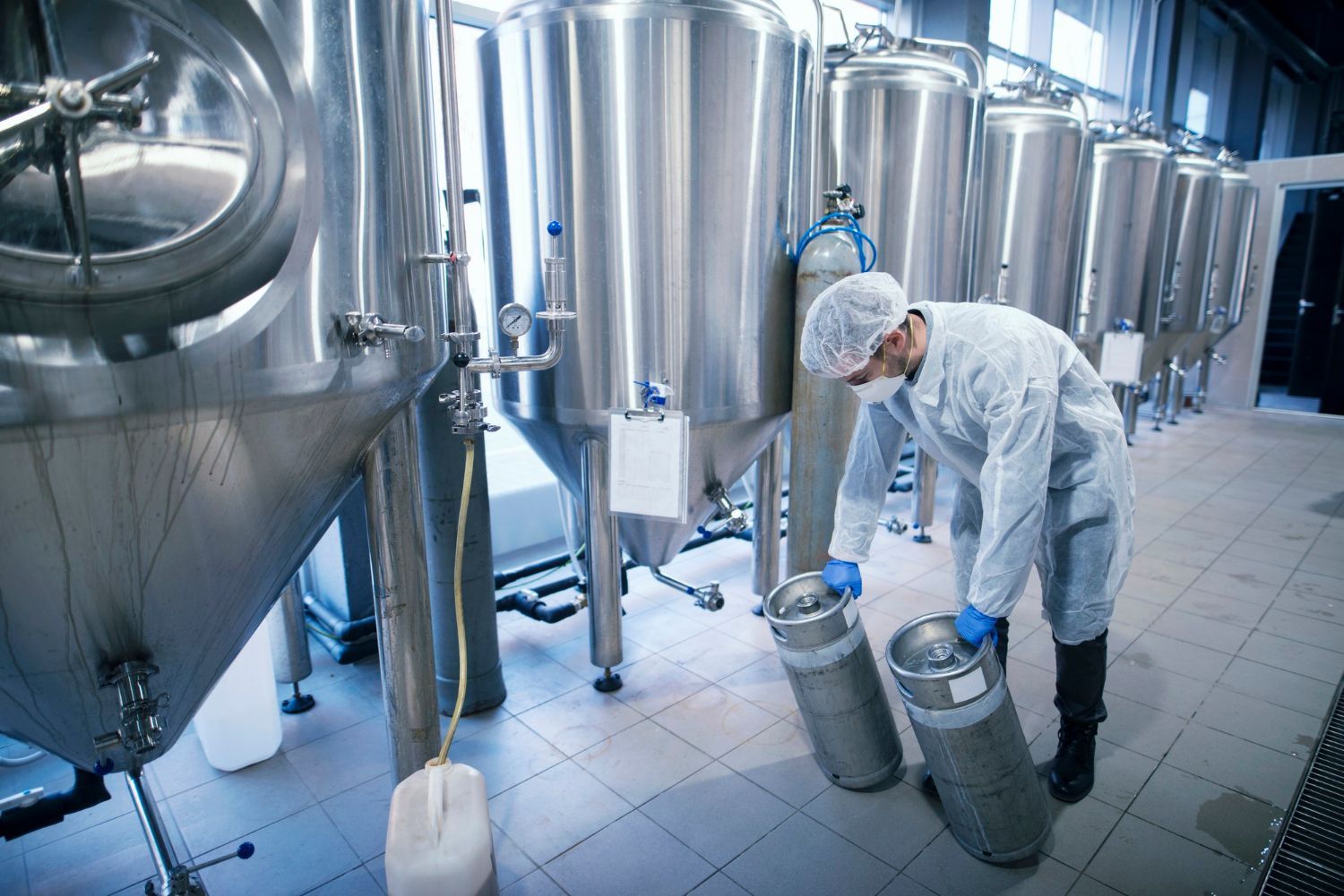 A worker in protective clothing and a hairnet moves stainless steel kegs in a facility with rows of large metal vats.