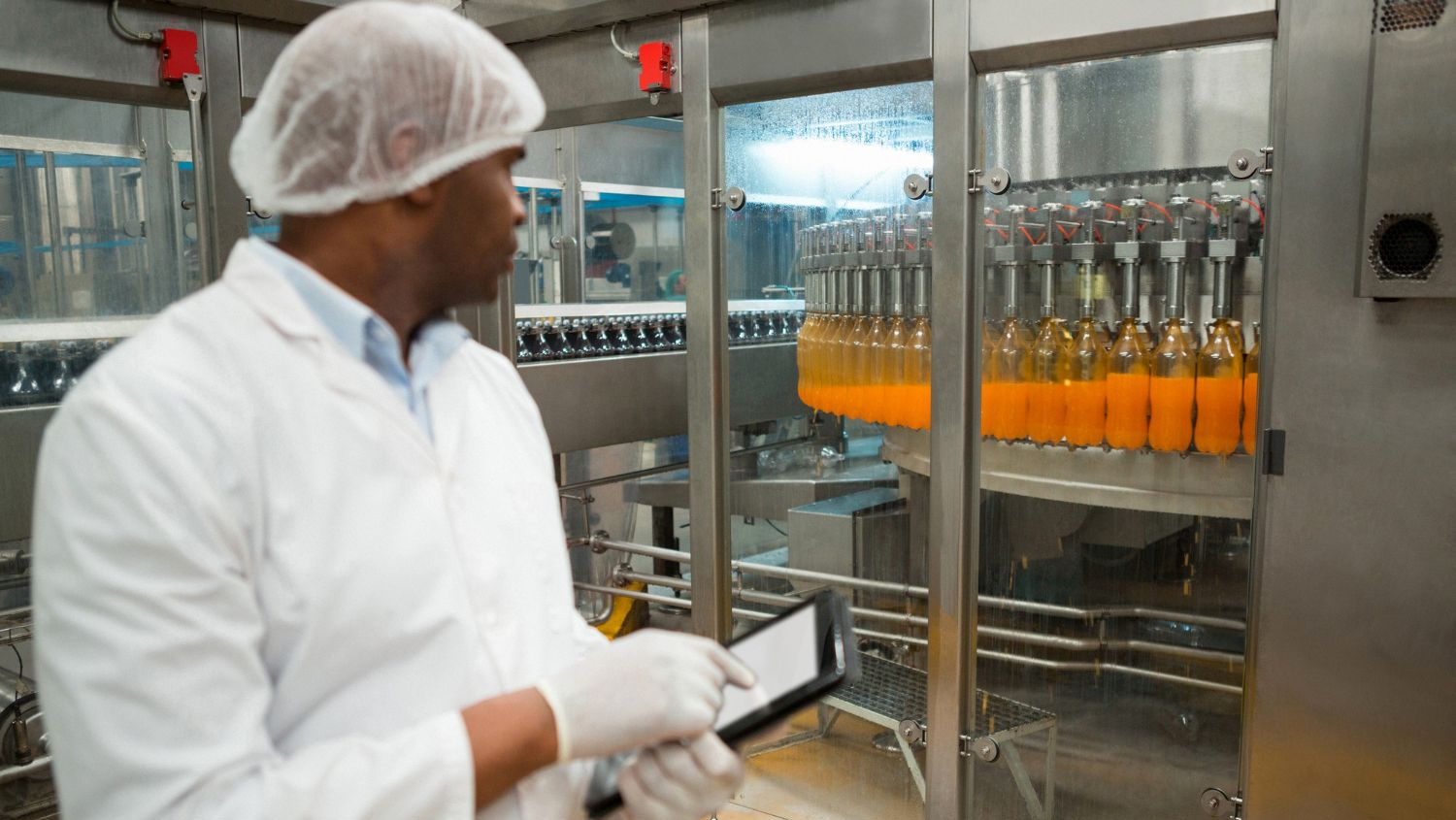 A worker in a lab coat, hairnet, and gloves holds a tablet while inspecting a line of orange-filled bottles in a factory.