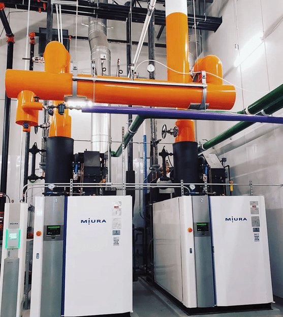 Two Miura boilers in a mechanical room, connected by bright orange insulated piping overhead.