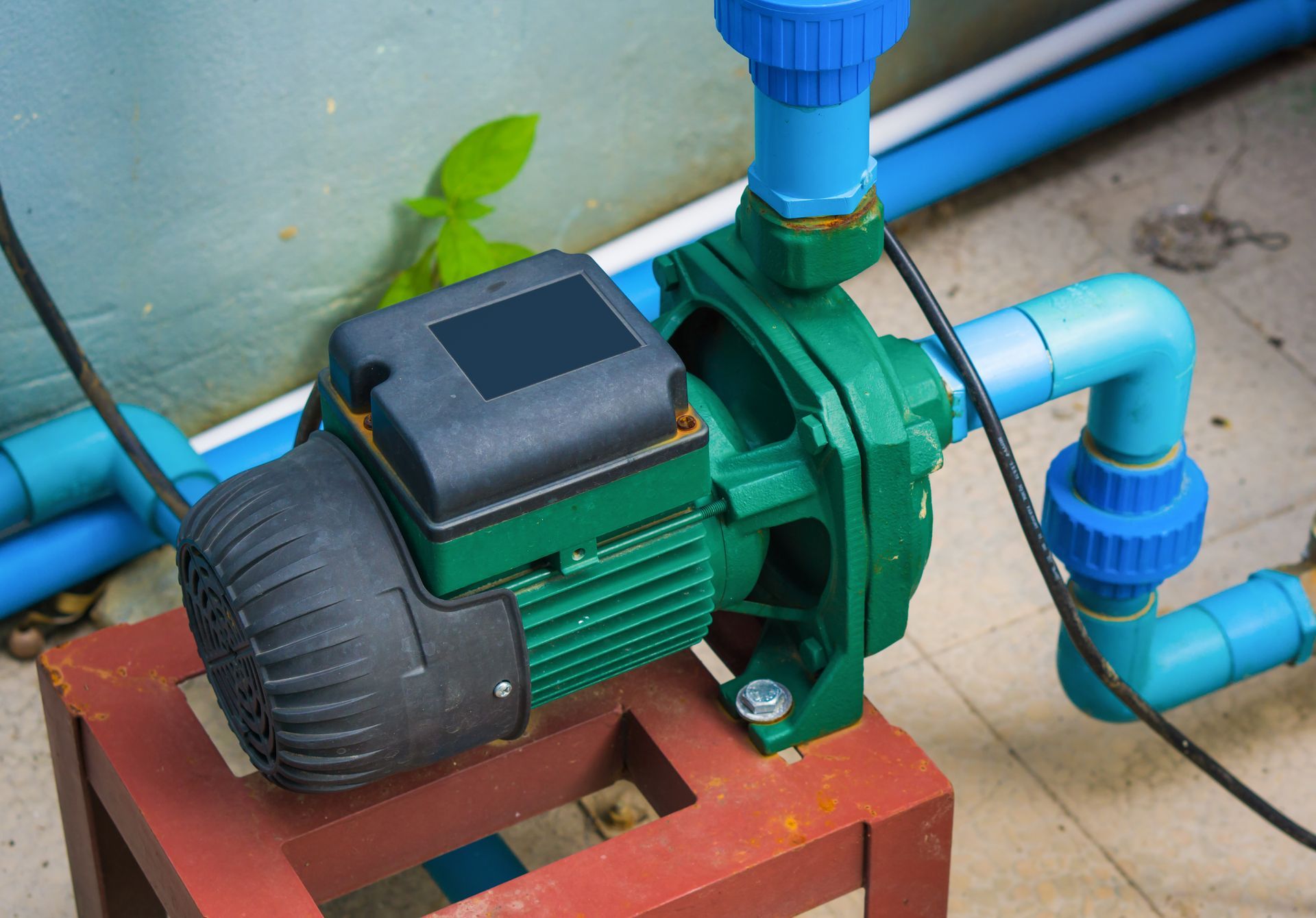 A green water pump is sitting on a wooden stool next to blue pipes.