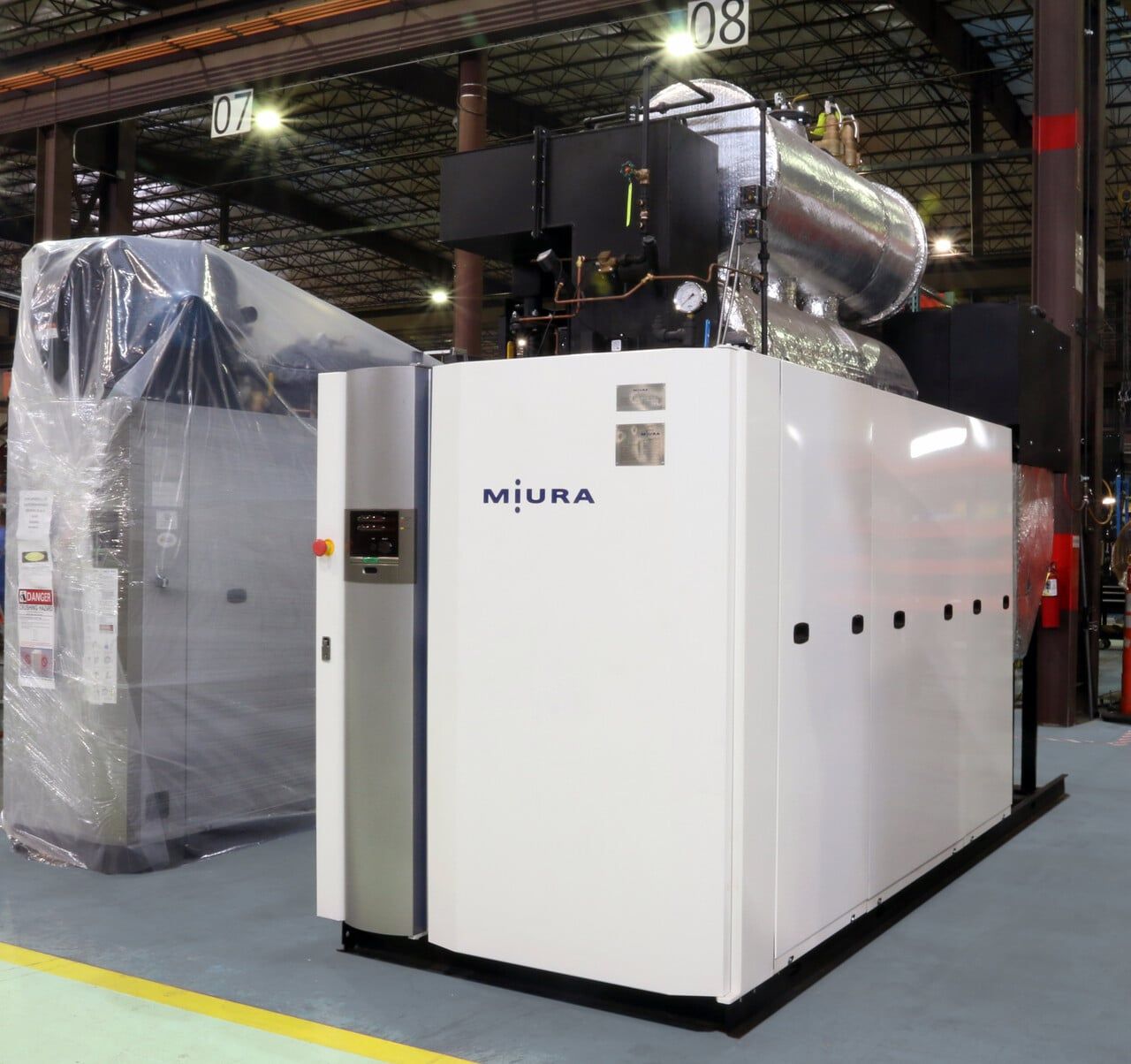 White industrial machine labeled “MIJUNA” in a factory, with a wrapped unit beside it.
