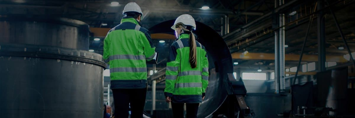 Two people wearing white hard hats and neon green high-visibility jackets stand in a factory, inspecting equipment.