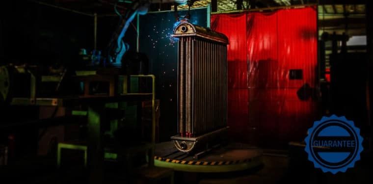 A tall, metallic radiator stands on a rotating platform in a dimly lit industrial workshop with a red curtain background.