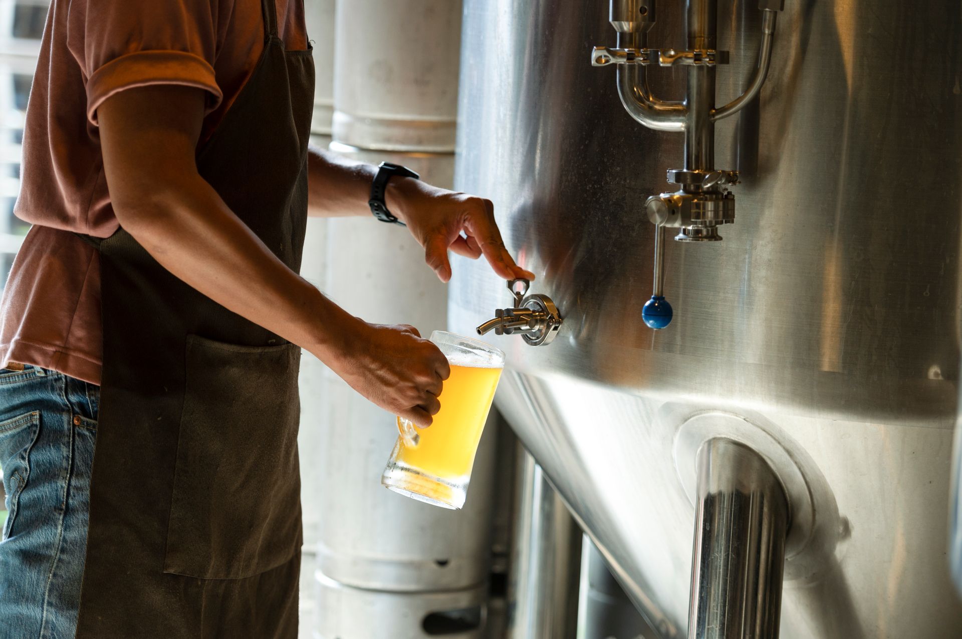 A person wearing an apron pours beer from a large industrial metal tank into a glass mug.