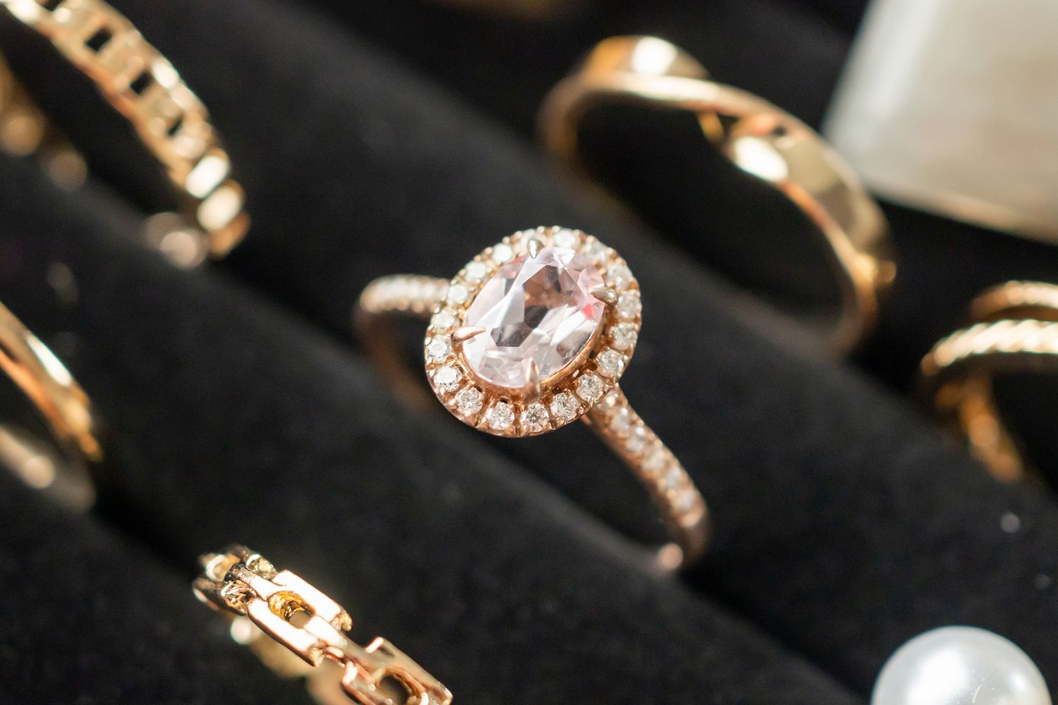 Close-up of a gold ring with an oval pink gemstone, surrounded by small diamonds, resting on black velvet, with other jewelry.