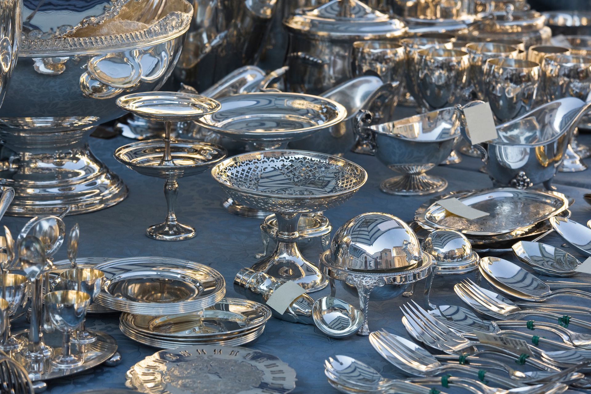 A table filled with shiny silver serving dishes and utensils, reflecting light. A table filled with shiny silver serving dishes and utensils, reflecting light.