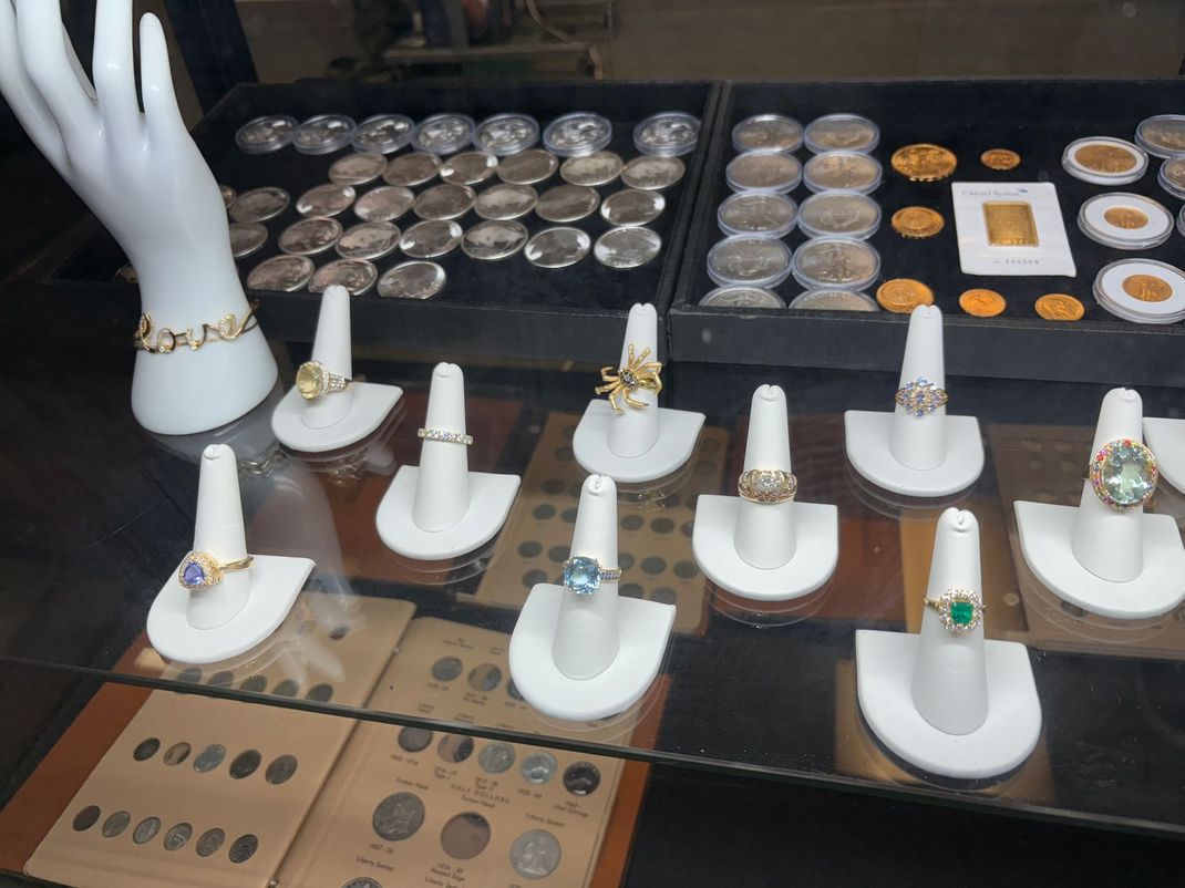 Woman with long wavy brown hair, wearing a gold sequin dress and red lipstick, looking down, and holding her hair; black background. Jewelry display with rings, bracelets, coins, and gold bar in a shop setting.