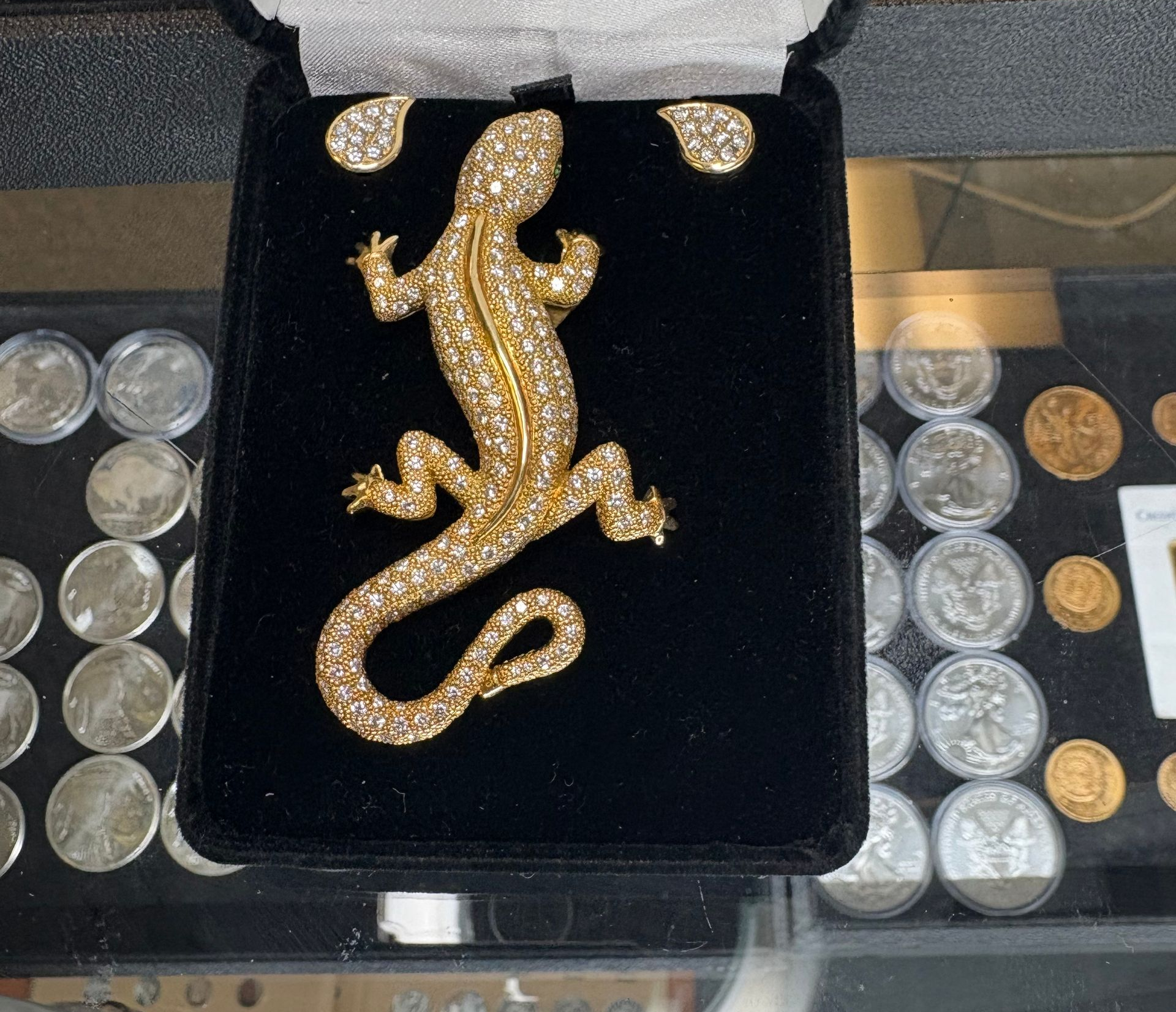 Gold jewelry, including a bracelet, earrings, and ring, encrusted with sparkling clear gemstones. Gold-colored jeweled lizard brooch in a black display case, coins in background.