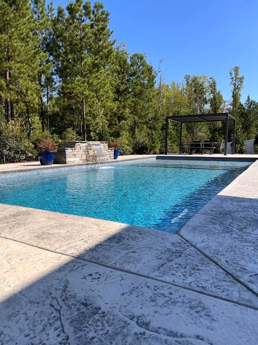 Outdoor swimming pool with blue water, stone deck, trees, and a shaded pergola under a clear sky