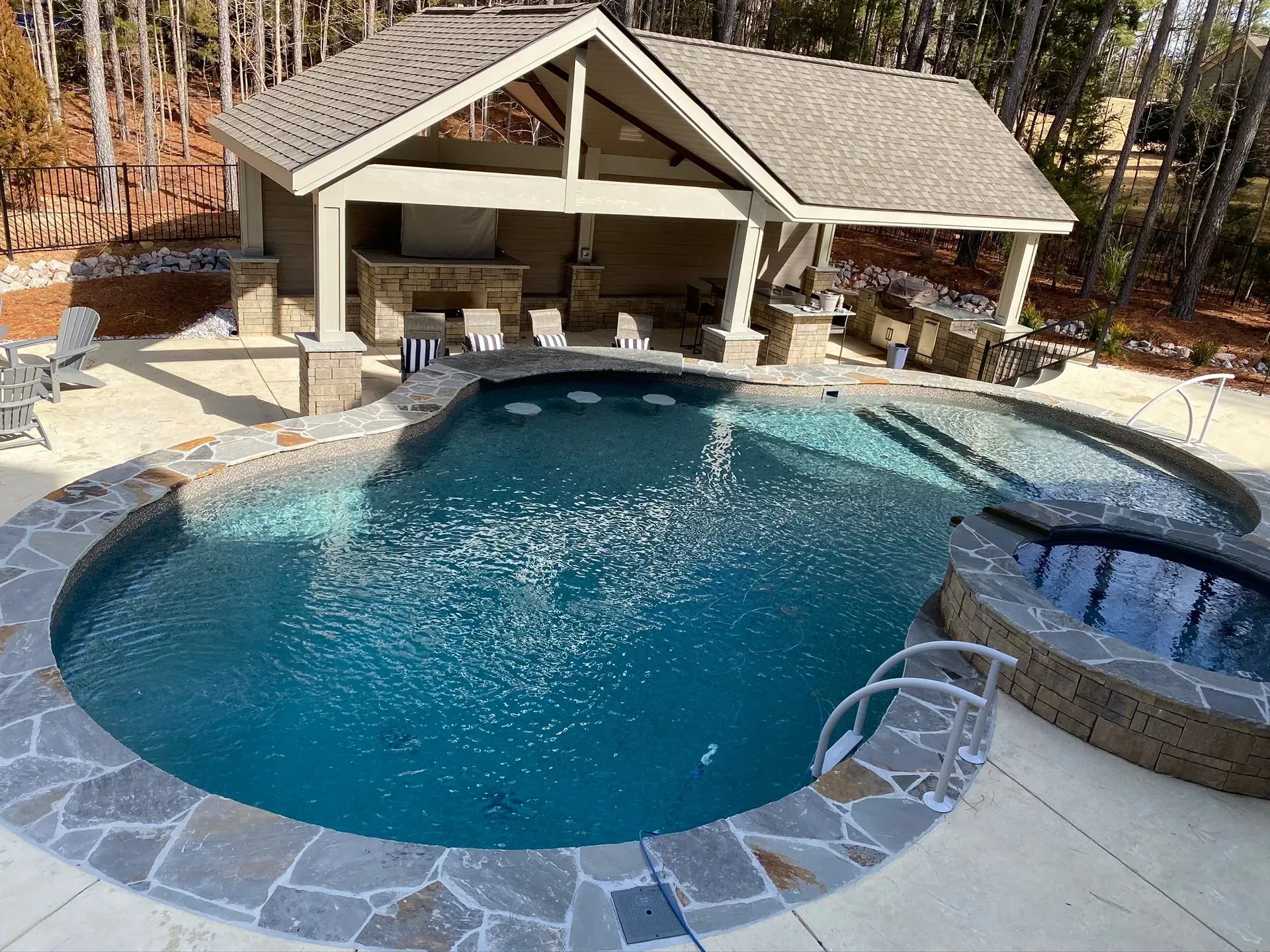 Backyard pool with stone deck, covered patio, and attached hot tub surrounded by trees