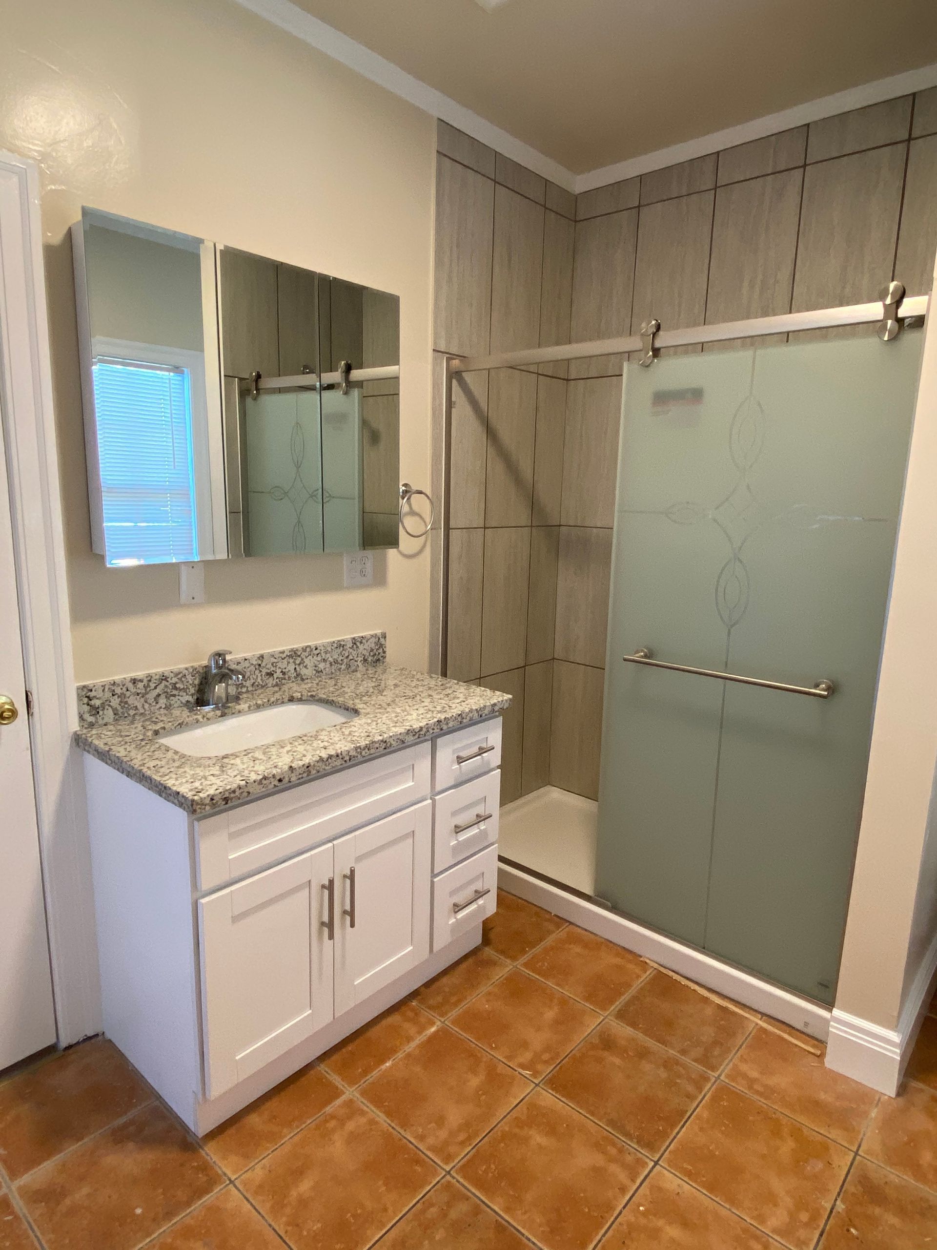 Bathroom with white vanity, granite countertop, mirror, and frosted glass shower enclosure on terracotta tile floor