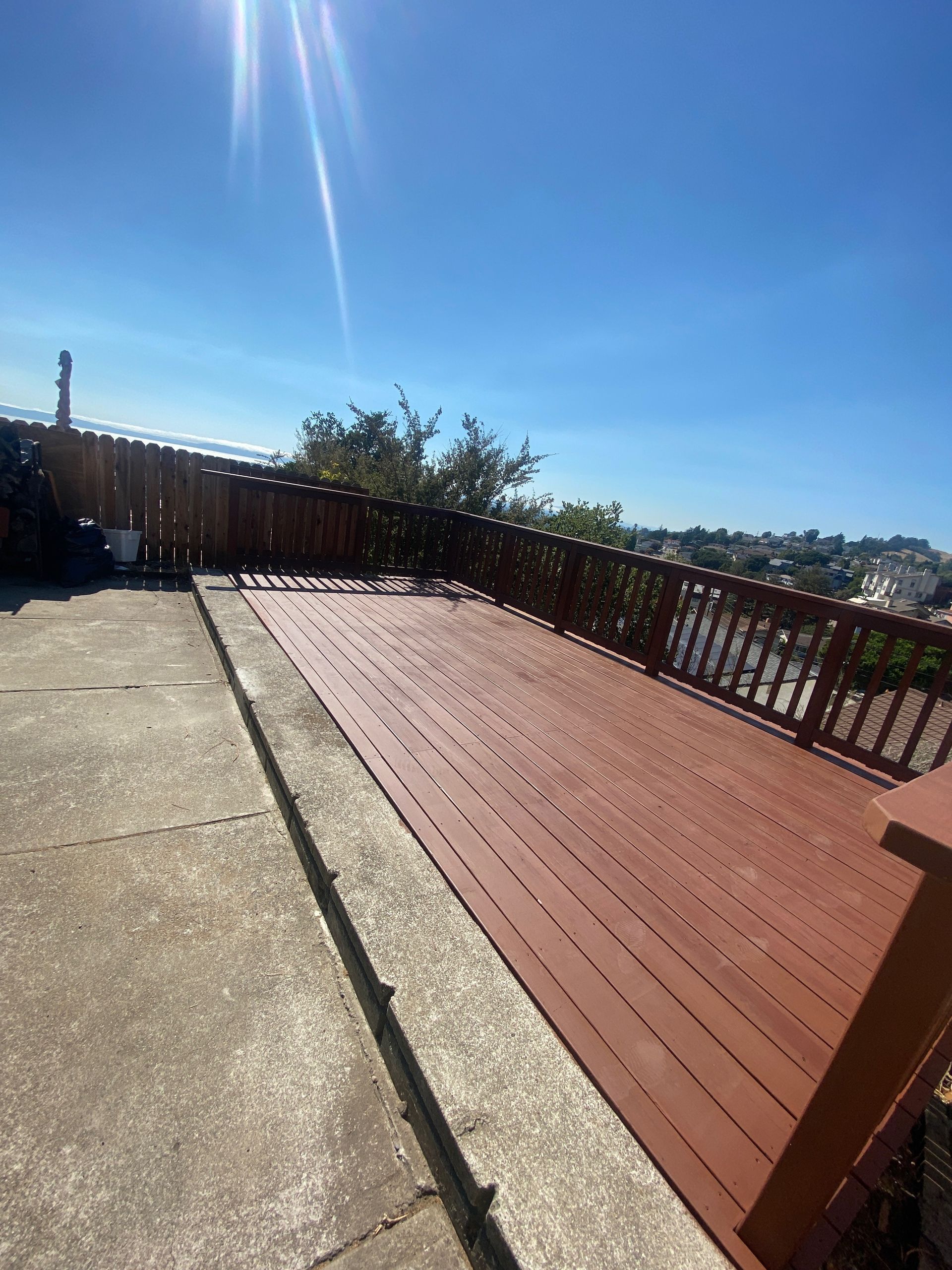 Sunny patio with concrete and red wooden deck, railing, and distant hillside view.