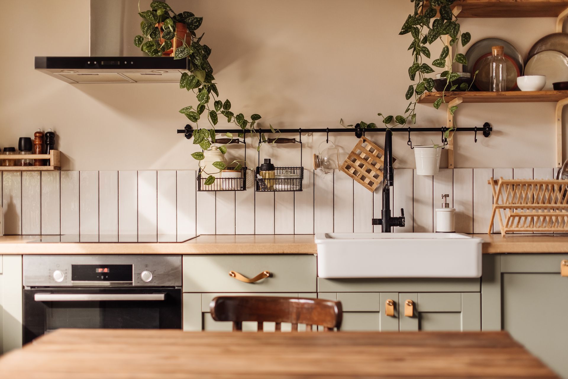 Cozy kitchen with sage cabinets, white farmhouse sink, wooden table, shelves, and trailing plants