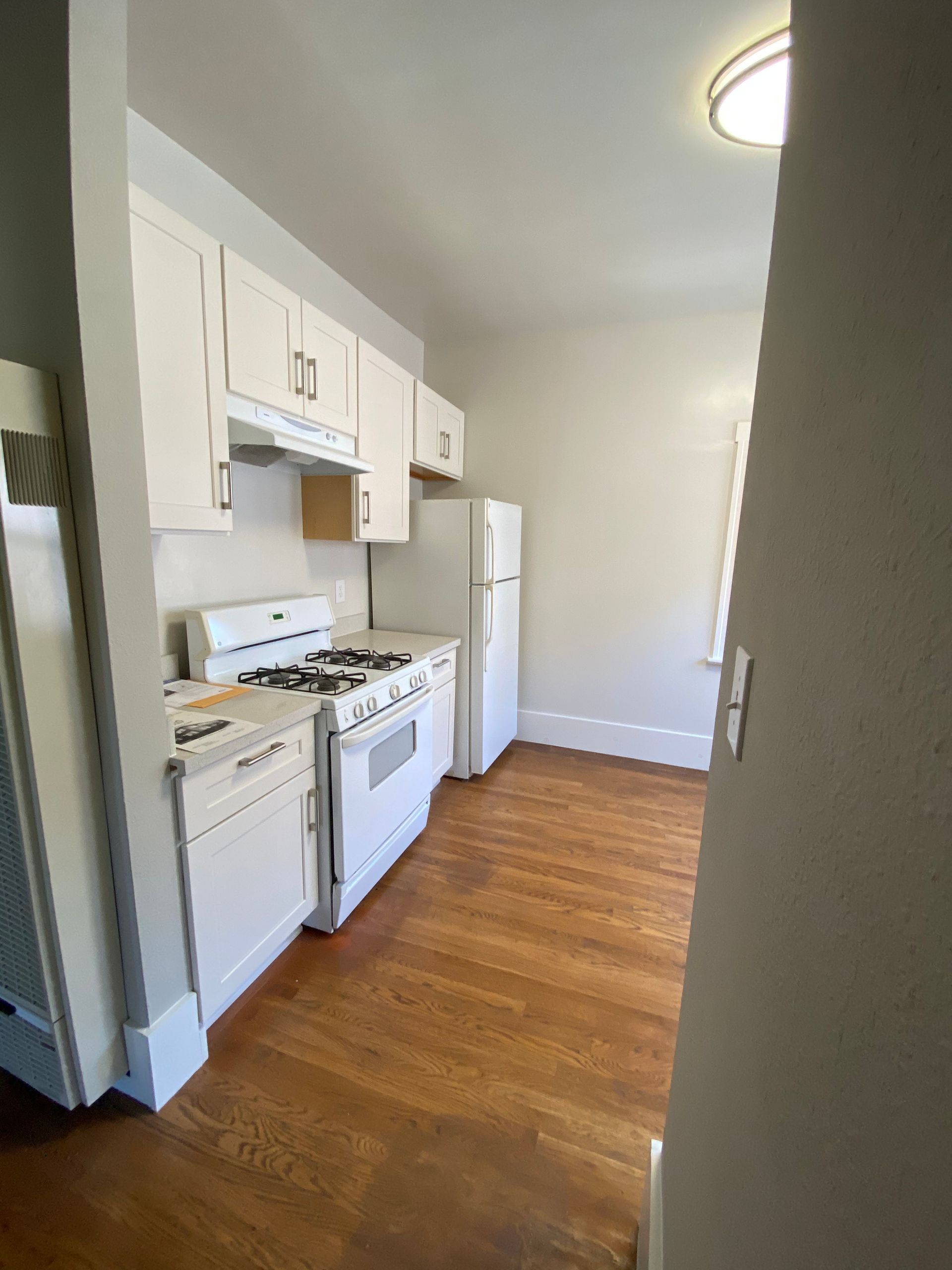 Small white kitchen with stove, refrigerator, and wood floor in a narrow apartment layout