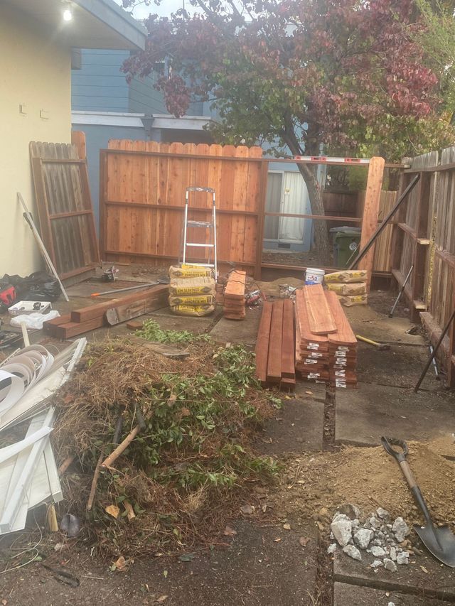 Outdoor construction yard with stacked lumber, a ladder, and fencing under a tree.