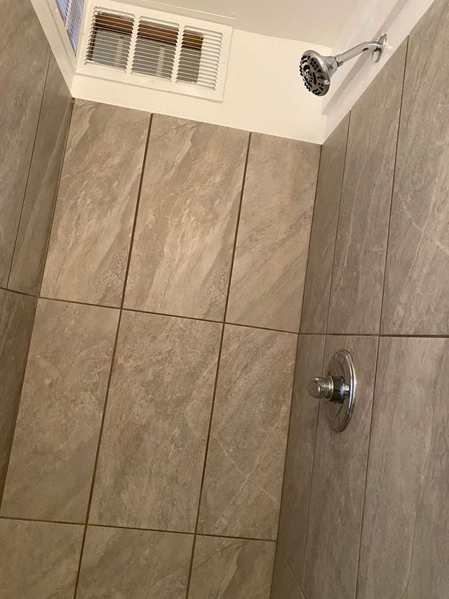Tiled shower wall with showerhead, knob, and vent in a bathroom.