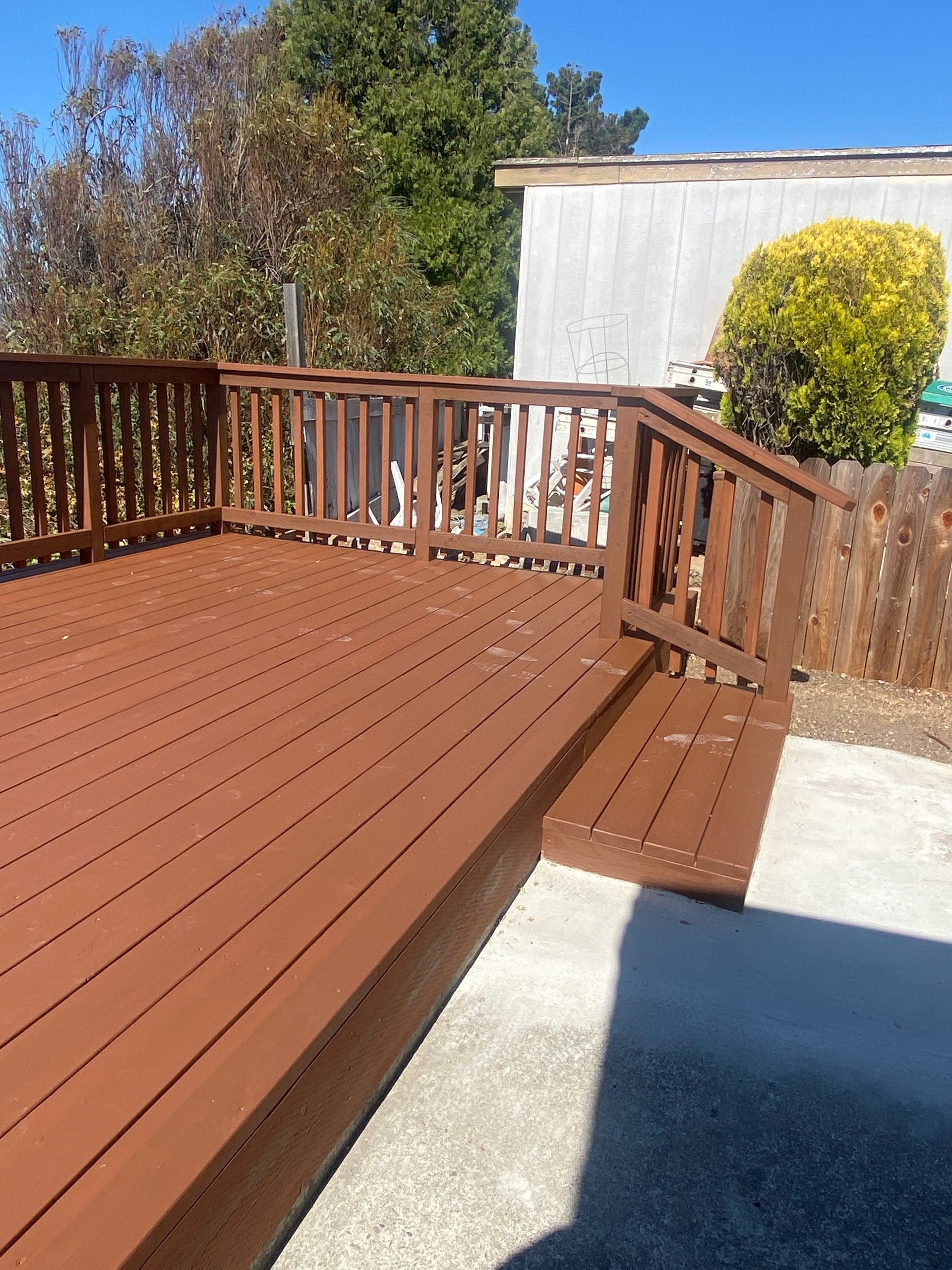 Wooden backyard deck with railings and a small step, beside a concrete patio and trees in sunlight