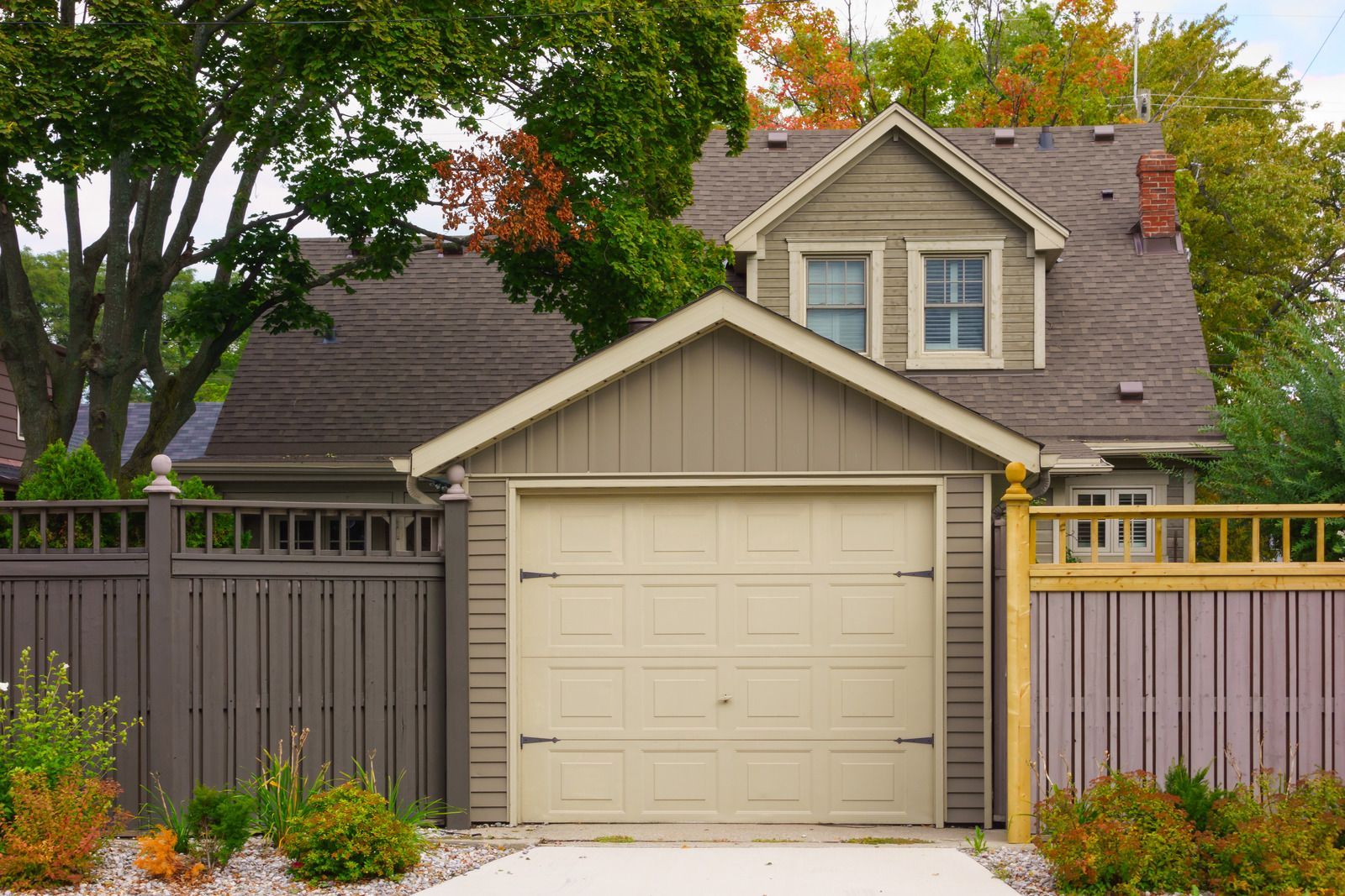 Suburban house with beige garage, gray siding, wooden fence, and leafy trees in front yard