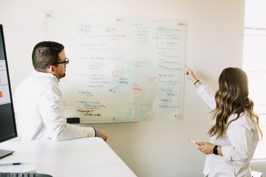 A man and a woman are standing in front of a whiteboard.