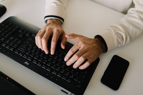 A person is typing on a keyboard next to a cell phone.