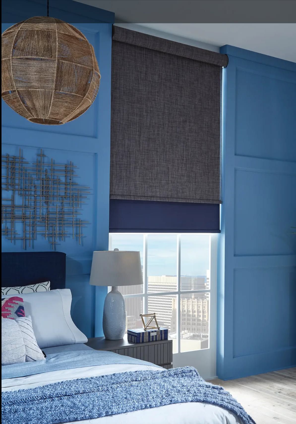 Blue bedroom with a bed, woven wall decor, and a window with a dark roller shade.