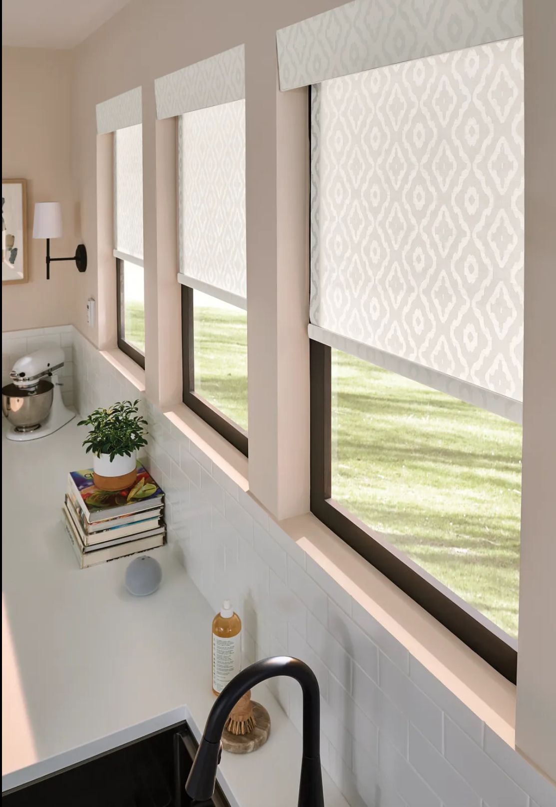 Bright kitchen sink beneath three beige roller shades over windows, with a potted plant on the counter.