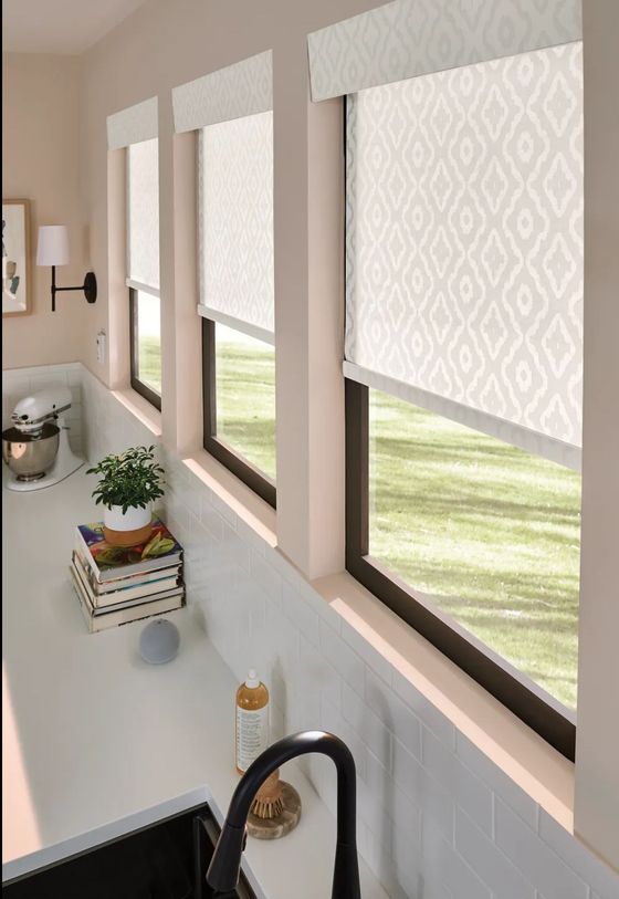 Kitchen window with three patterned roller shades, sunlight, sink faucet, and a small plant on the counter