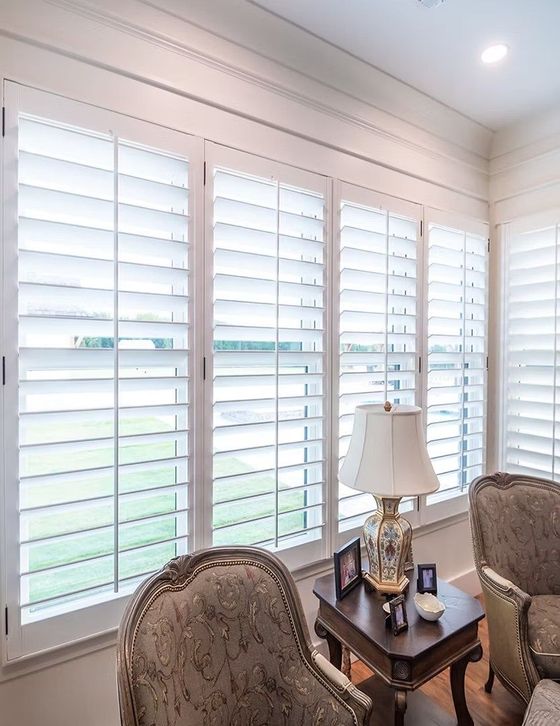 Bright sitting room with white plantation shutters, upholstered chairs, and a side table with a lamp.