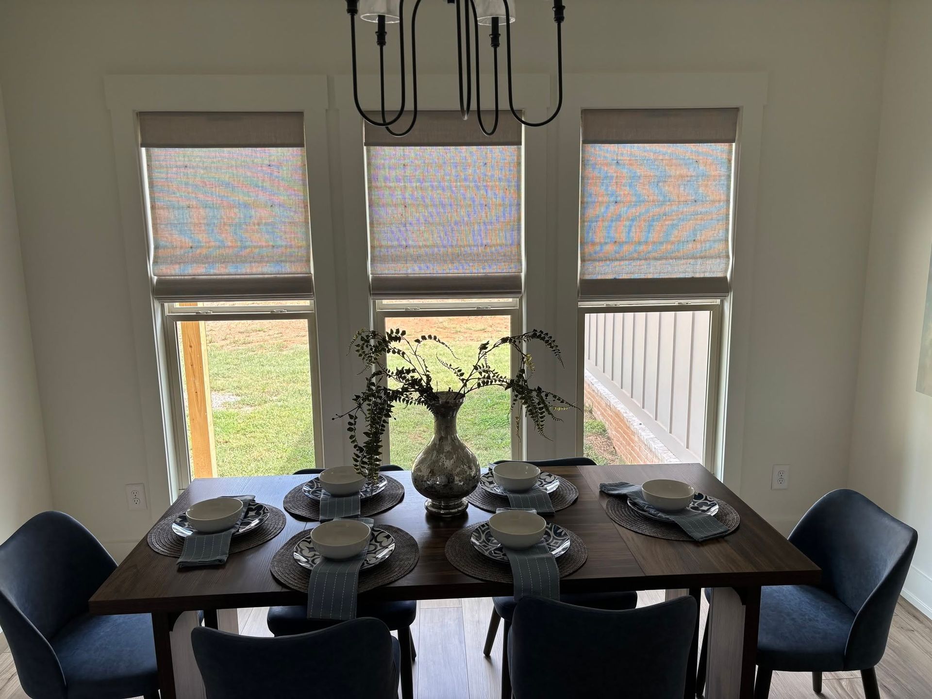 Dining table set for six beneath three windows, with dark chairs, white dishes, and a floral centerpiece.