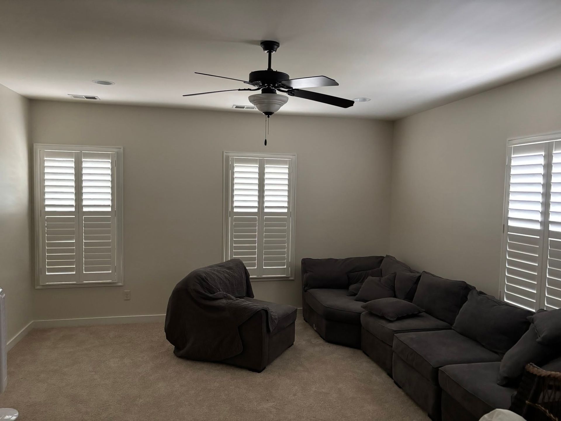 Empty living room with gray sectional sofa, recliner, ceiling fan, and three windows with white shutters.