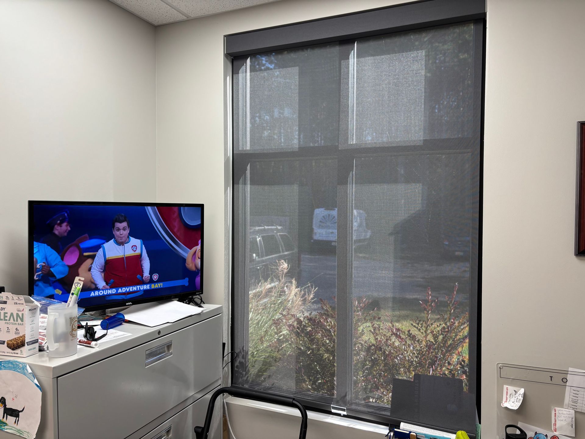 Office corner with a TV on a cabinet and a large window with a dark roller shade.