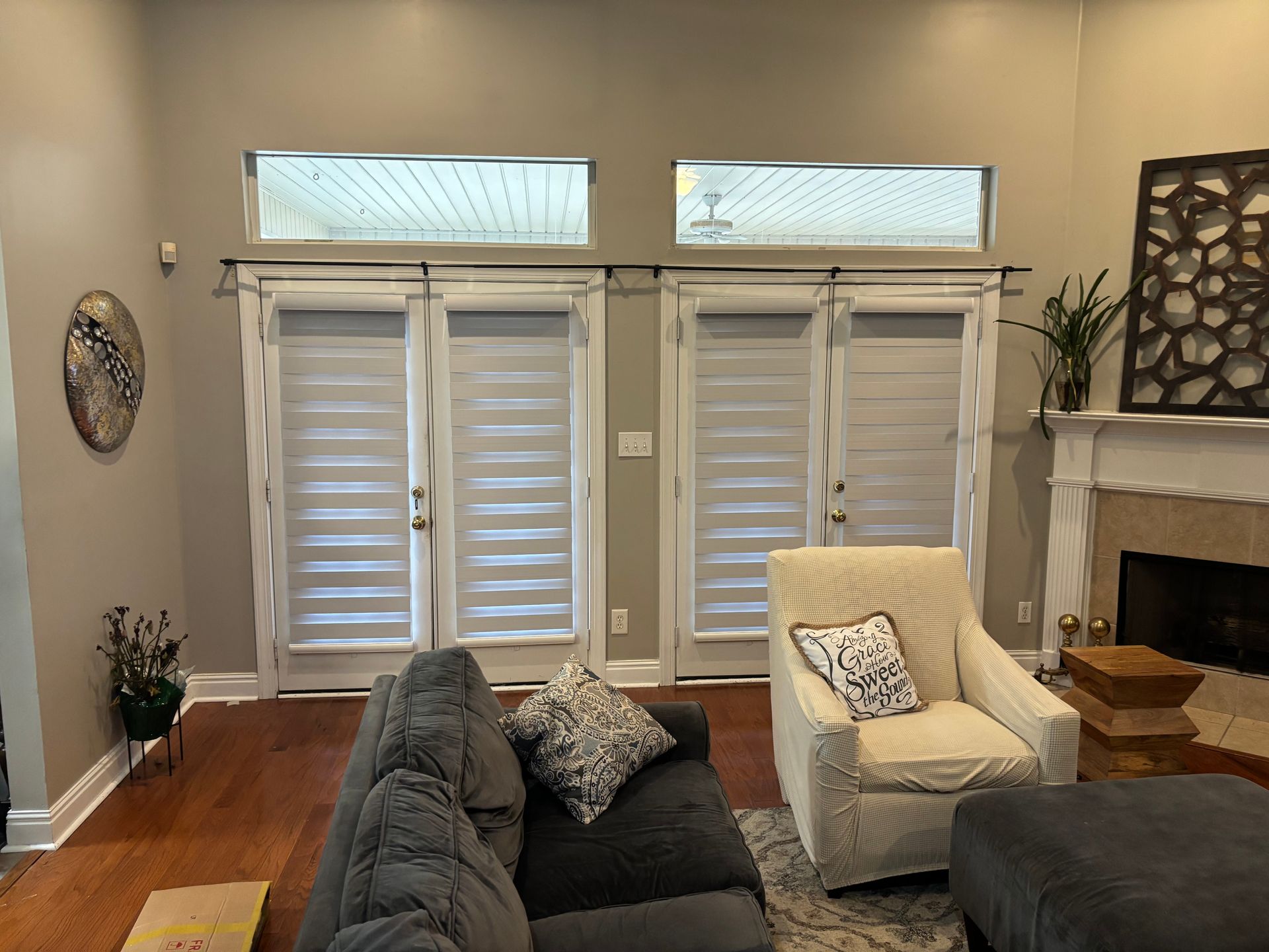 Bright living room with white shutters, beige walls, a dark sofa, cream chair, and fireplace.