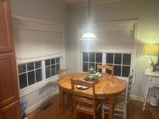 Dining nook with round wooden table, chairs, pendant light, and two windows with blinds