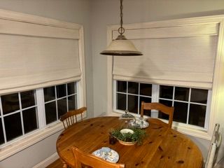 Round wooden dining table by two windows with blinds and a hanging pendant light.