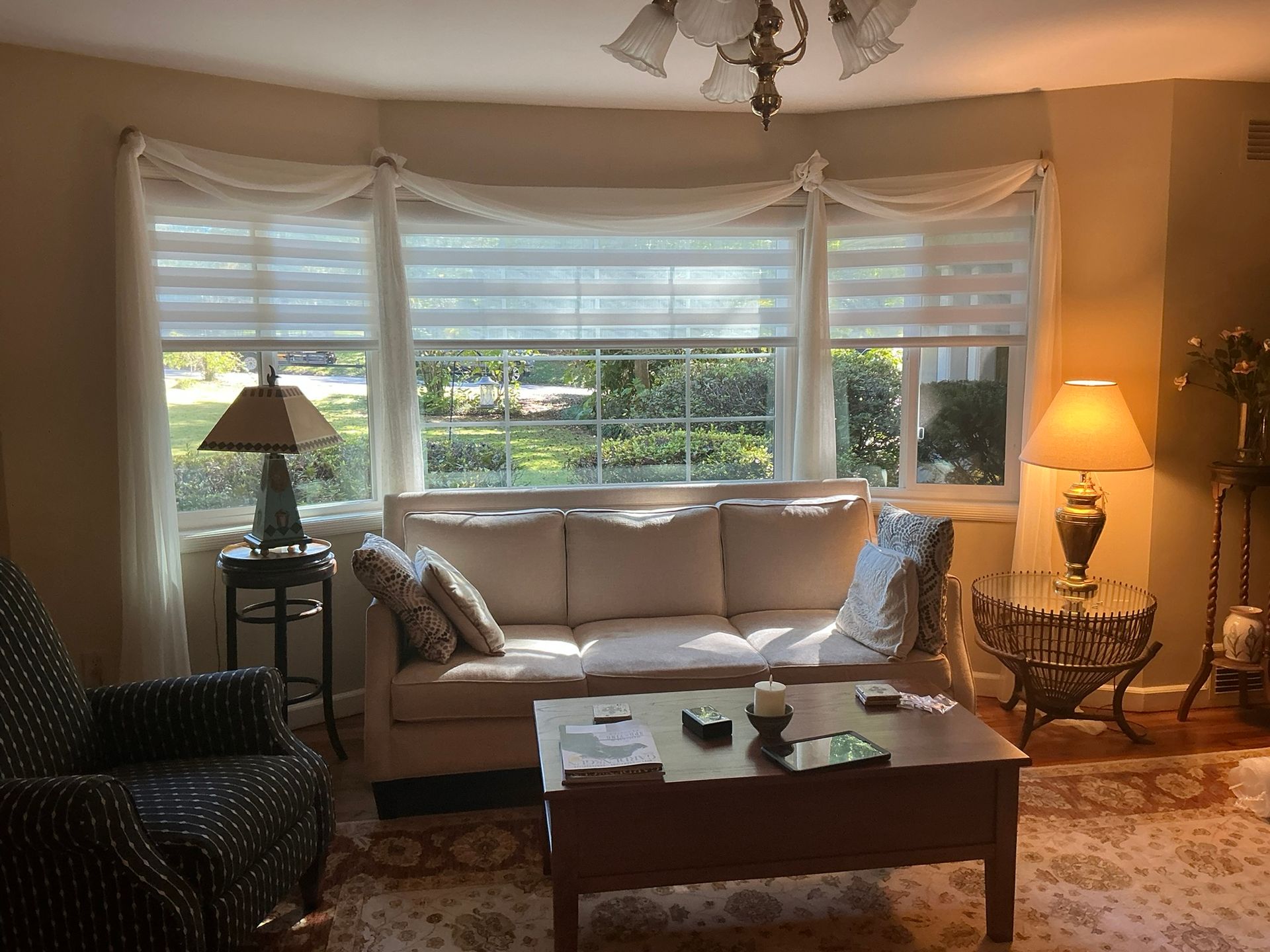 Cozy living room with beige sofa, coffee table, lamps, and large sunlit window with sheer drapes