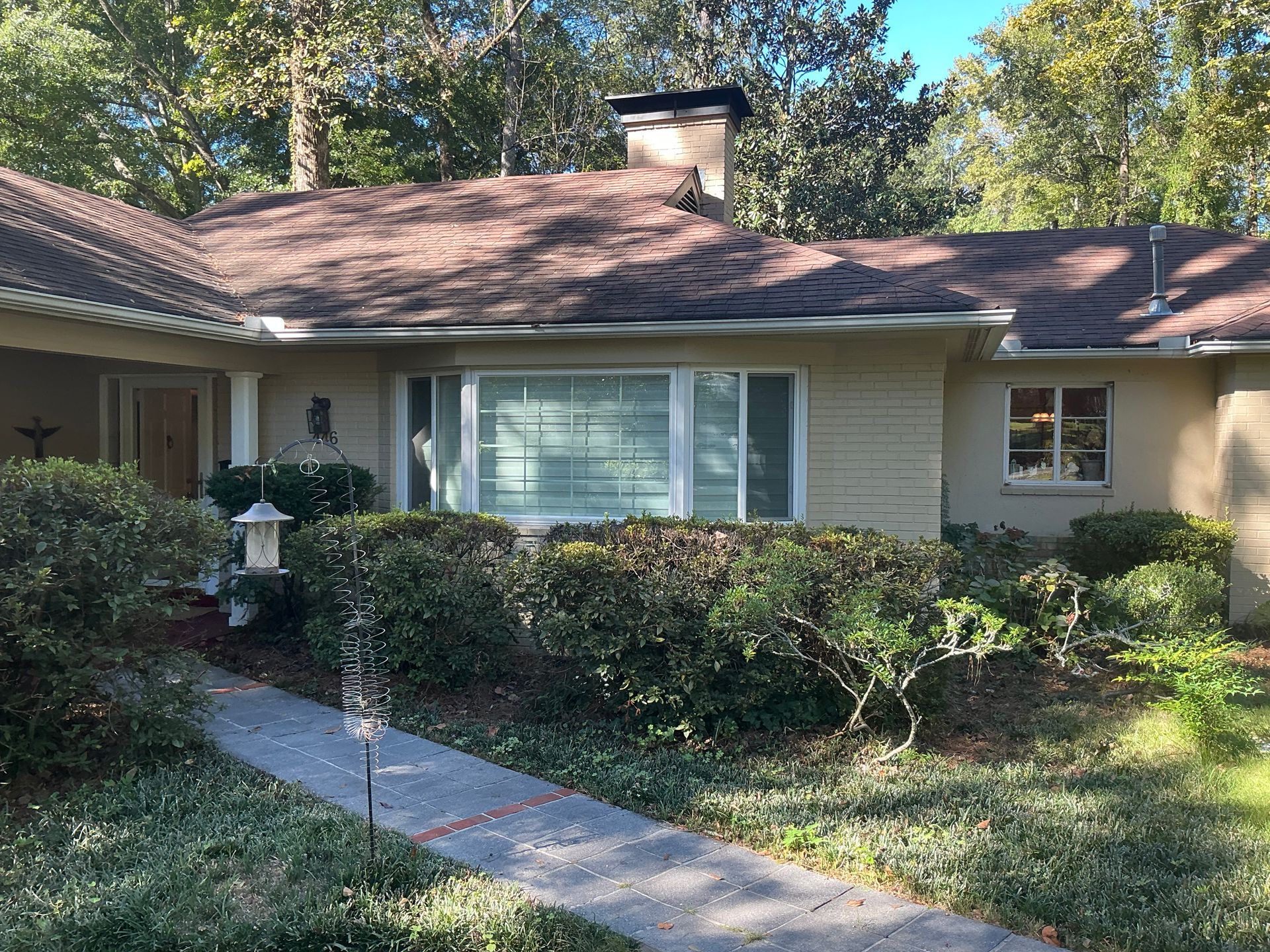 Single-story beige house with white shutters, front walkway, and bushes in a sunny yard