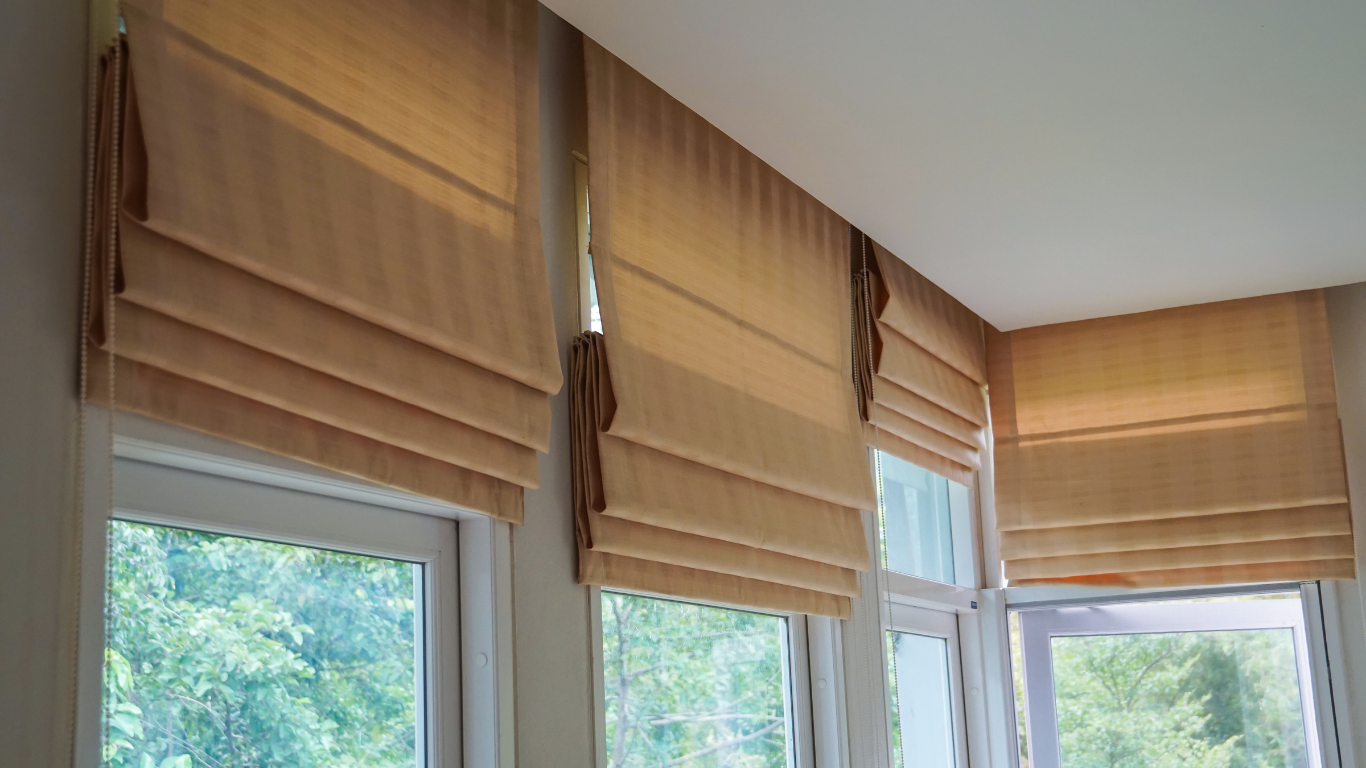 Beige Roman shades covering corner windows with greenery visible outside