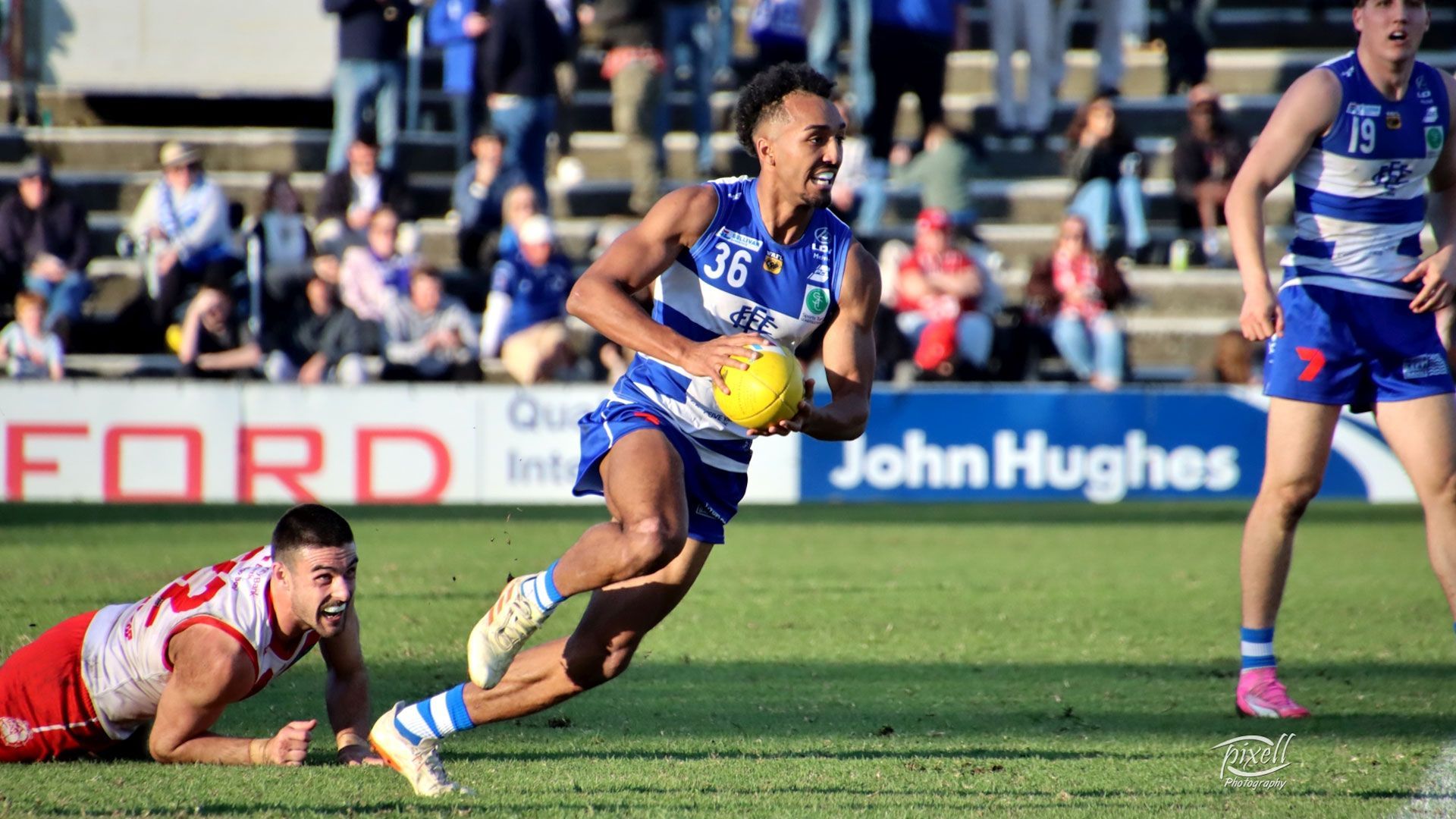 Player in blue and white uniform runs with ball, other player lunging on the ground. Green field, crowd in background.