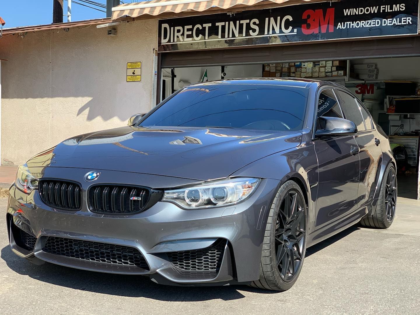 Dark gray BMW M3 parked in front of Direct Tints Inc. window tint shop, with tinted windows.
