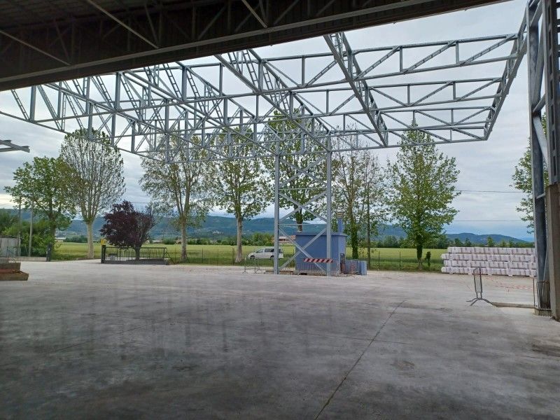 Padiglione a cielo aperto con tetto a traliccio in acciaio, pavimento in cemento e alberi che si affacciano sui campi circostanti.