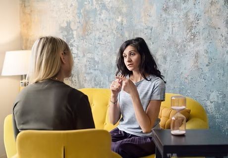 Two women are sitting on a yellow couch talking to each other.