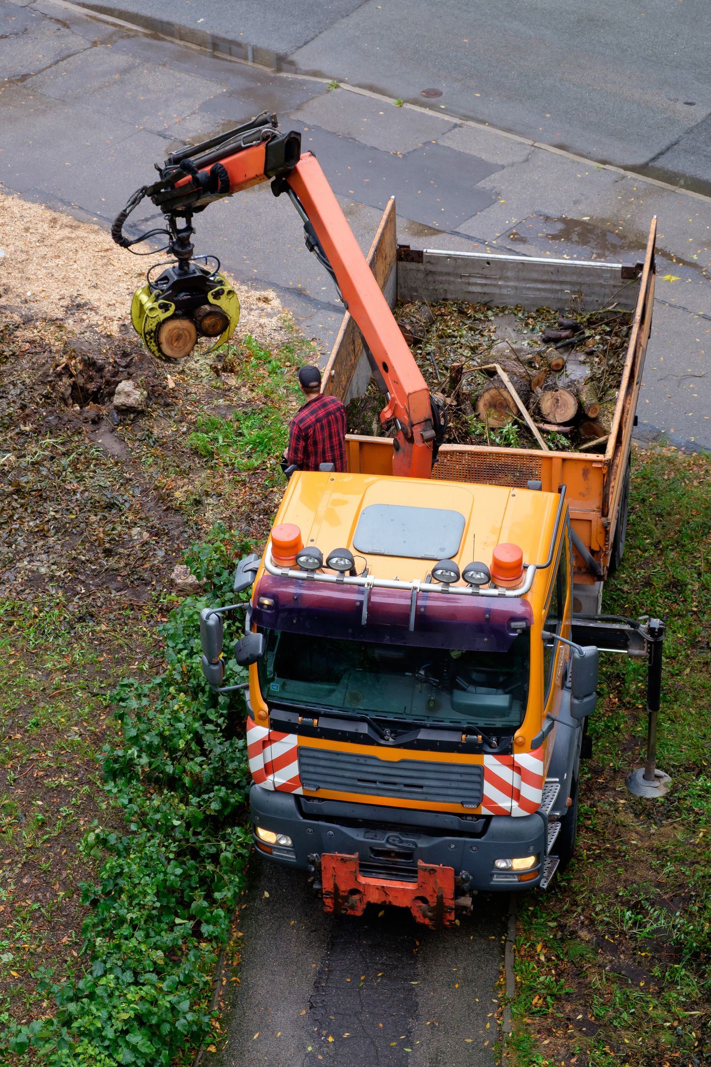 Yellow truck with crane loading debris from a tree stump into its bed.