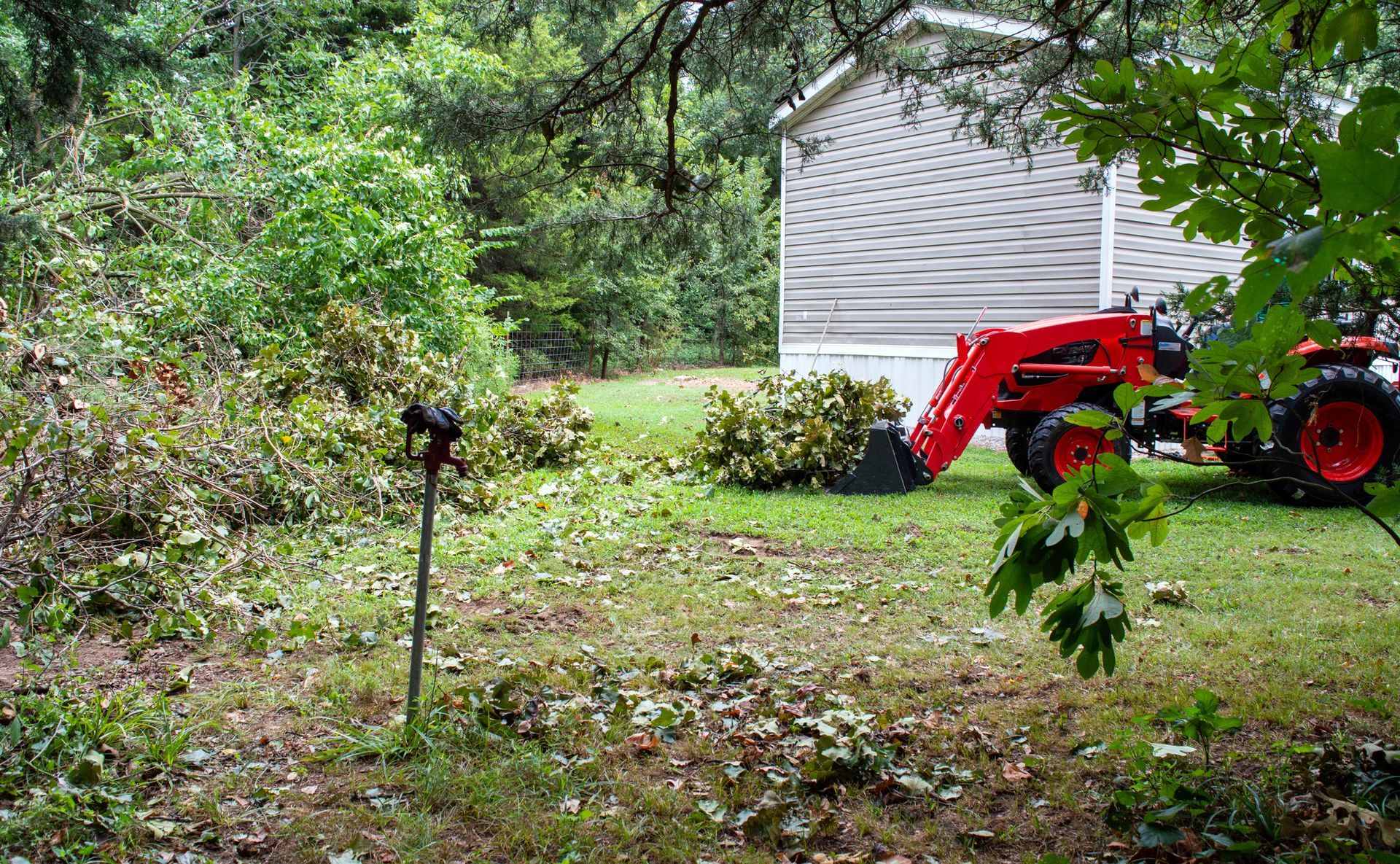 Orange tractor with front loader clearing brush next to a building; overcast day.
