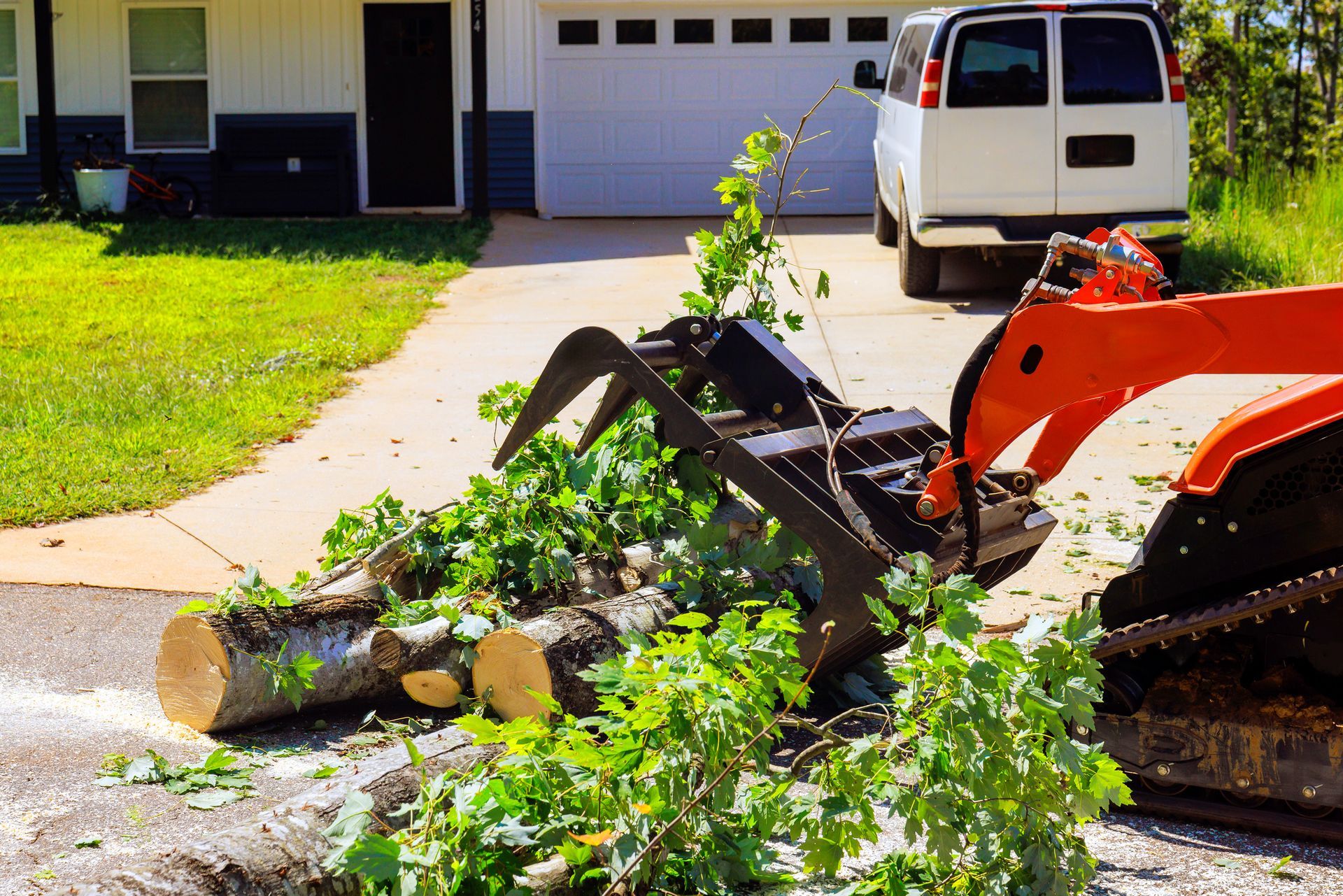 Orange and black grapple truck holding logs and branches on a driveway near a white van.