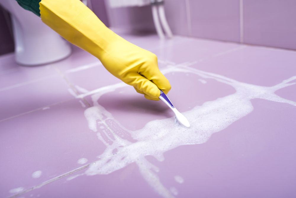 A Person Wearing Yellow Gloves is Cleaning a Bathroom Floor With a Toothbrush — Cleaning for Good in Port Macquarie, NSW