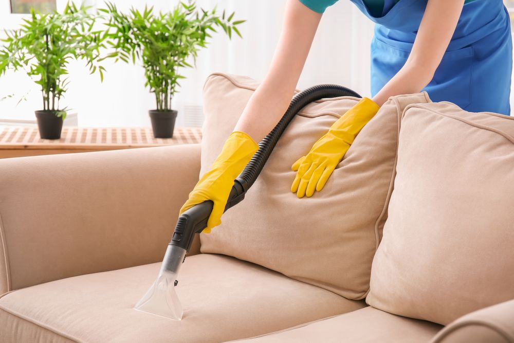 Person using a vacuum cleaner to clean a beige couch, wearing yellow gloves, indoors — Cleaning for Good in Byron Bay, NSW