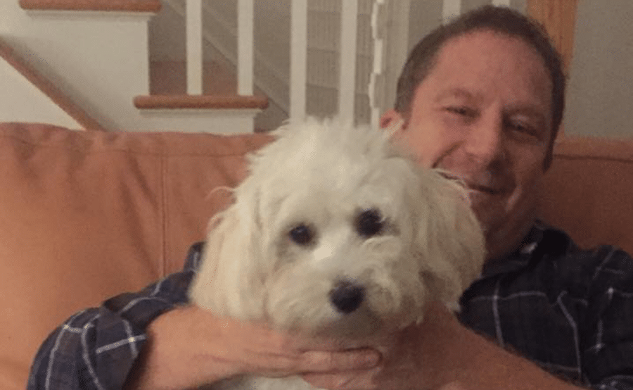 A man is sitting on a couch holding a small white dog.