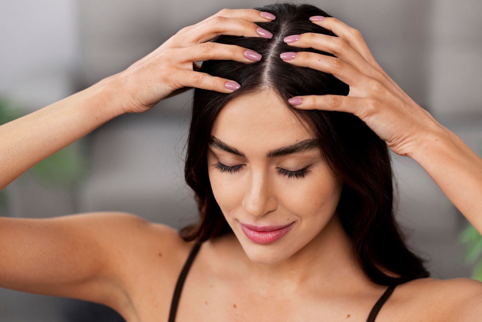 Woman massaging her scalp with both hands, looking down against a blurred indoor background